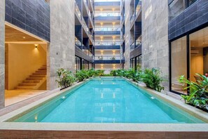 A modern indoor swimming pool with a central staircase, surrounded by plants and with a high ceiling. - Modern 1BR Condo | Rooftop Pool & Gym, Near Beach (Playa del Carmen)