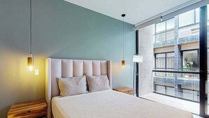 A modern bedroom with a large bed, two bedside tables, and a view of a building through a large window. - Modern 1BR Condo | Rooftop Pool & Gym, Near Beach (Playa del Carmen)