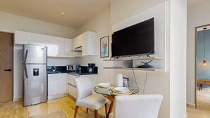 A modern kitchen with white cabinets, a built-in refrigerator, a microwave, and a coffee maker. There is a dining area with a glass table and white chairs. A flat-screen TV is mounted on the wall above the dining area. - Modern 1BR Condo | Rooftop Pool & Gym, Near Beach (Playa del Carmen)