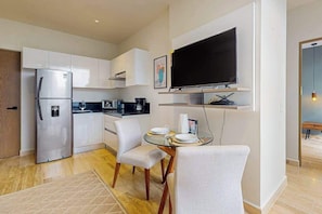 A modern kitchen with white cabinets, a built-in refrigerator, a microwave, and a coffee maker. There is a dining area with a glass table and white chairs. A flat-screen TV is mounted on the wall above the dining area. - Modern 1BR Condo | Rooftop Pool & Gym, Near Beach (Playa del Carmen)
