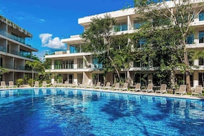 A swimming pool with lounge chairs, surrounded by multi-story buildings with balconies and trees. - Coastal 2BR Playacar Condo - Workspace & Pool (Playa del Carmen)