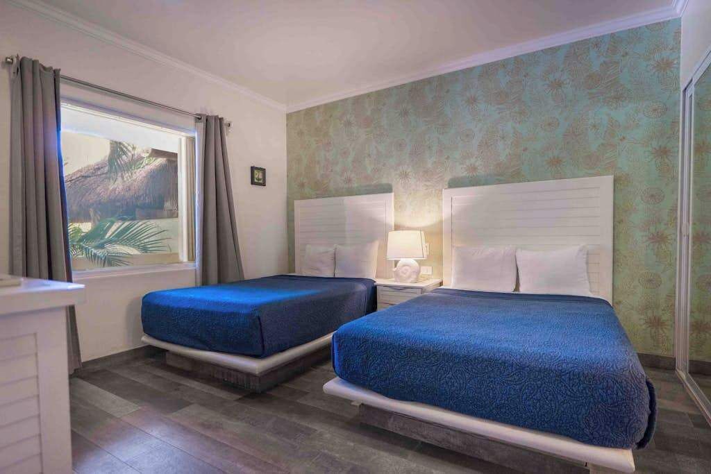 Une chambre d’hôtel avec deux lits, une fenêtre avec des rideaux, une table de chevet avec une lampe et un mur décoré d’un motif.