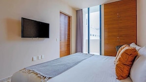 A hotel room with a bed, a TV mounted on the wall, a window with curtains, and a wooden wardrobe. - Playa Studio | Rooftop Pool & Balcony (Playa del Carmen)