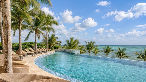 Une piscine à bords incurvés, entourée de chaises longues et de palmiers, avec vue sur l’océan.