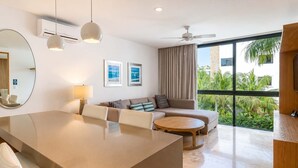Une chambre d’hôtel moderne dotée d’un coin repas, d’un canapé, d’une table basse ronde, d’un ventilateur de plafond et de grandes fenêtres offrant une vue sur la verdure.