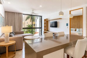 A modern hotel room with a sofa, a round coffee table, a dining table with chairs, a TV mounted on the wall, and a large window with a view of the outside. - Modern Playa del Carmen Apt: Rooftop Pool & WiFi (Playa del Carmen)