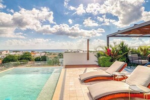 Una piscina en la azotea con sillones de descanso, una pérgola y vistas al mar y al paisaje urbano.