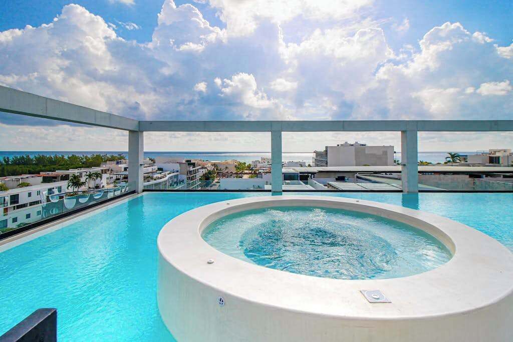 A rooftop infinity pool with a hot tub, surrounded by a white structure, overlooking a coastal cityscape.