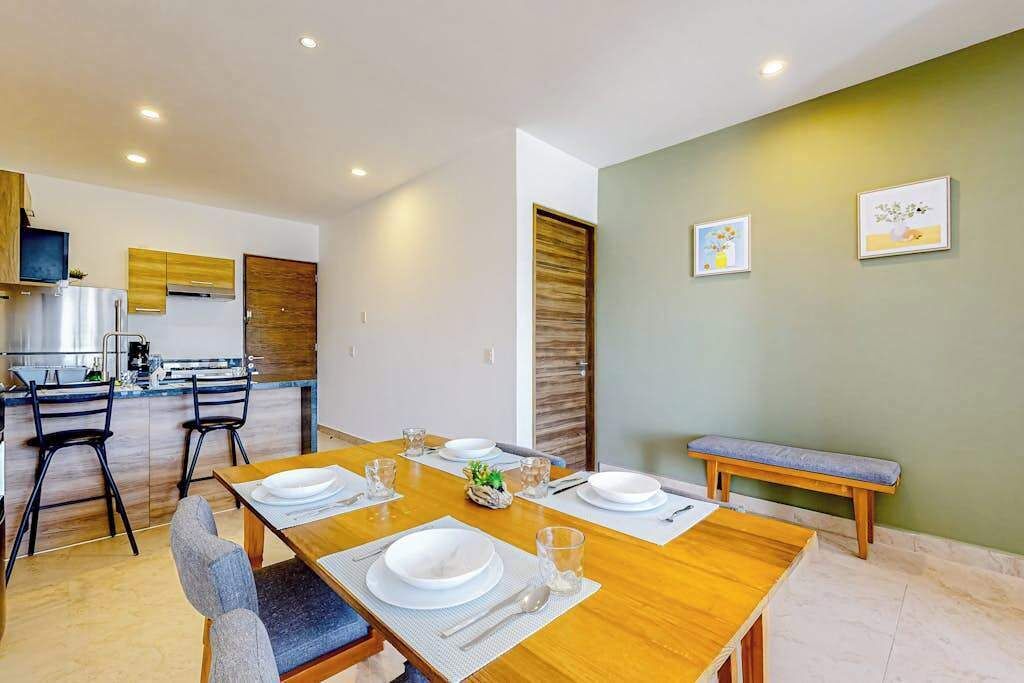 A dining area with a wooden table set for four, a kitchen in the background, and a bench against the wall.