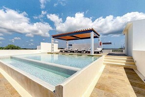 A rectangular swimming pool with a pergola and lounge chairs, surrounded by a white building and a clear sky. - 1BR condo near Akumal beach, rooftop pool (Akumal)