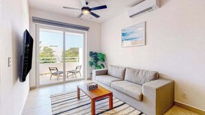 A living room with a sofa, a coffee table, a TV, a ceiling fan, and a large window. - 1BR condo near Akumal beach, rooftop pool (Akumal)