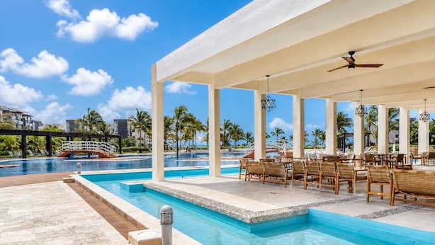 A poolside area with a covered seating area, a bridge, and palm trees in the background. - Beachfront 3BR Condo, Resort Pool | Playa (Playa del Carmen)