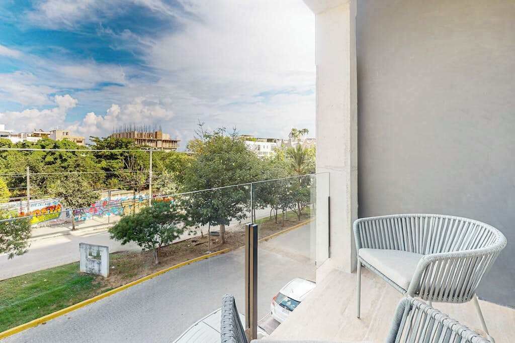 Een balkon met twee witte rieten stoelen, uitzicht op een straat met bomen en gebouwen, en een heldere lucht.