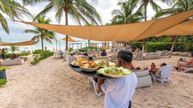 Un área de comedor frente a la playa, con mesas y sillas bajo una toldilla, palmeras y vista al océano.