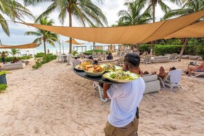 A beachside dining area with tables and chairs under a canopy, palm trees, and a view of the ocean.
