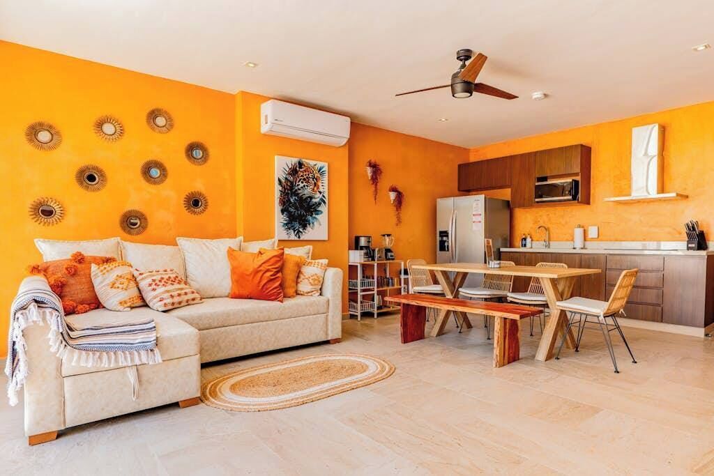 A living room with a sofa, coffee table, and dining area with chairs. There is a kitchen area with cabinets and a refrigerator.