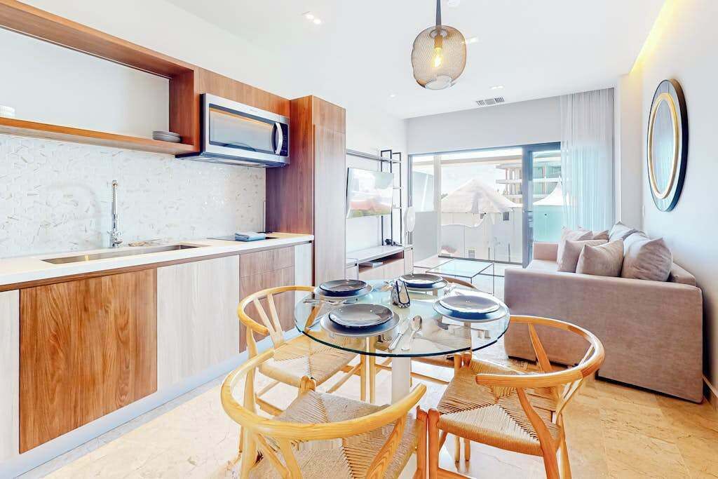 A modern kitchen with a built-in microwave, a dining area with a glass table and wicker chairs, and a living area with a sofa and a large mirror.