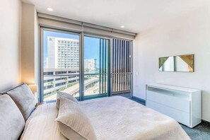 A modern hotel room with a large window, a bed, a white dresser, and a city view. - Ocean view condo w/ luxury pool rooftop access (Cancún)