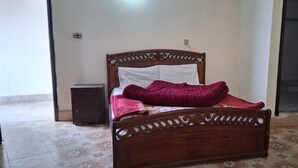 Una cama de madera con cabecera, una manta roja y un mueble auxiliar al lado.