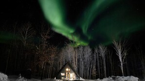 Een chalet met verlicht interieur, omringd door sneeuw en bomen, onder een nachthemel met zichtbare poollichten.