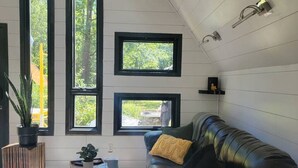 Een kamer met een schuine balkenkap, een zwart leren bank, een kamerkleed met dessin, en drie ramen met uitzicht op groen.