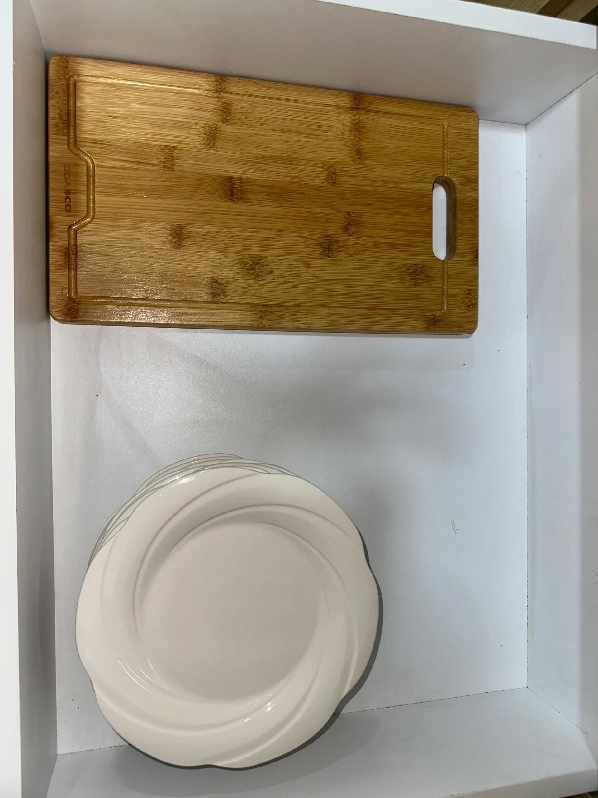 Un cajón blanco con una tabla de cortar de madera y una pila de platos blancos.