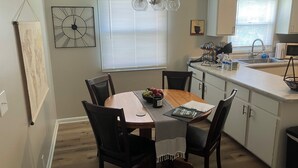 Una cocina con mesa, sillas, un reloj, una ventana con persianas y un lavamanos.