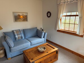 A blue sofa with a plaid throw, a wooden coffee table with a basket, a framed picture on the wall, a window with curtains, and a wreath. - serene wooded 3-bedroom house West Tisbury deck, screen porch and outdoor shower (West Tisbury)