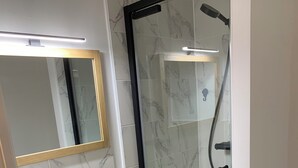 Ein Badezimmer mit einer in Glas eingeschlossenen Dusche, einem weißen Waschbecken mit Holzschubladen und einem Spiegel mit goldener Rahmung.