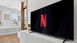 Ein modernes Wohnzimmer mit einem großen Flachbildfernseher, der an einem weißen Schrank befestigt ist, einem Fenster mit Blick auf ein Gebäude und einem Heizkörper unterhalb des Fensters.