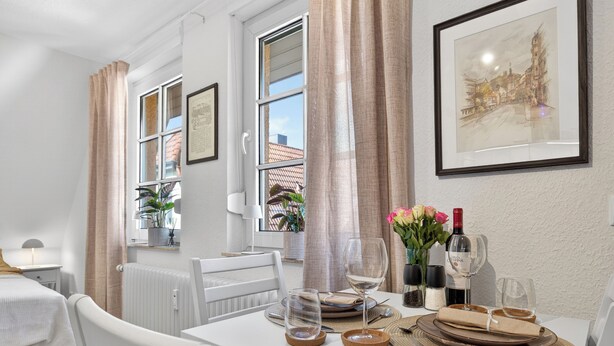 Ein ordentlich eingerichteter Essbereich mit einem Tisch für zwei Personen, einem gerahmten Kunstwerk an der Wand und einem Fenster mit Vorhängen.