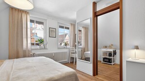 Ein Schlafzimmer mit Bett, Fenster, Heizkörper, Stuhl, Spiegel, einem Regal mit Küchengeräten und einer Lampe.