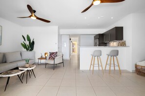 Ein modernes Wohnzimmer mit einer Couch, einem Couchtisch und Sesseln, einer Küchenzeile mit Barhocker und einem Deckenventilator.