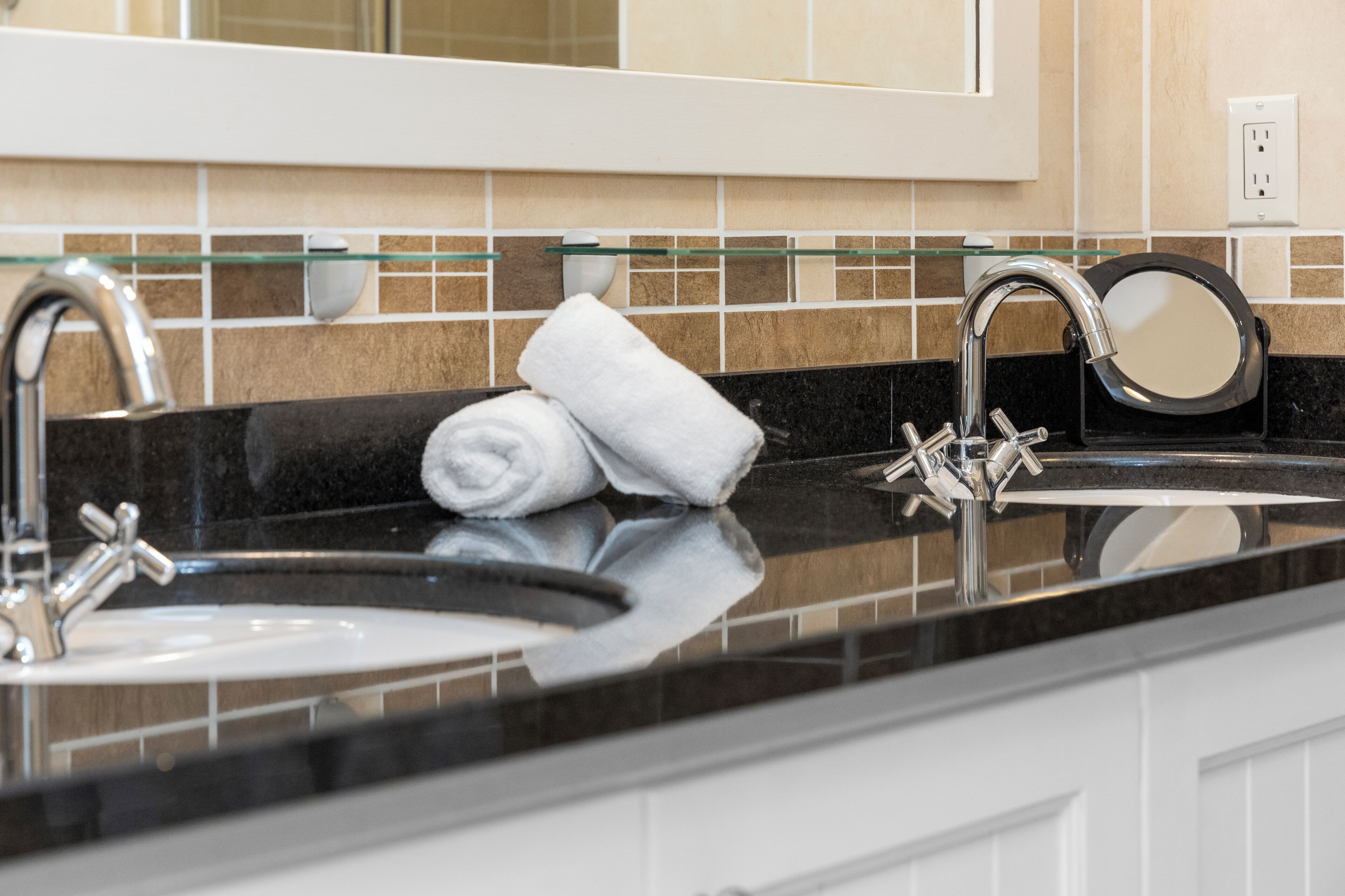 A bathroom with a black countertop, two sinks, a glass shelf, and a mirror.