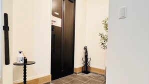 Een hotelgang met een zwarte deur, een klein tafeltje met een fles en een verpakking, een plant en een betegelde vloer met een rookvrij sign.