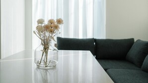 Een glazen vaas met bloemen op een witte tafel in een kamer met een donkergroene bank en transparante gordijnen.