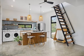 A modern kitchen with a central island, two bar stools, a washing machine, and a large window. - Casa Rocas Surf House 50m to the Beach! (Hacienda Iguana)