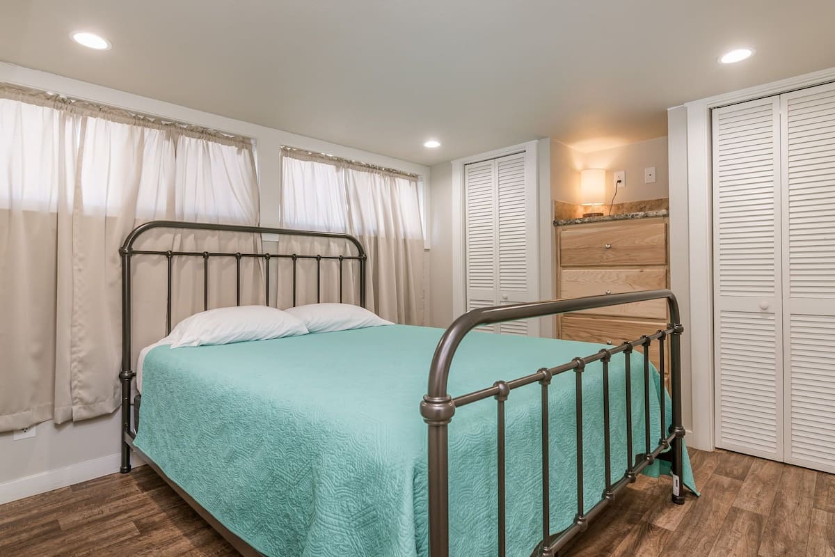 A single bed with a metal frame, a teal bedspread, and white pillows. There is a wooden dresser with a mirror, a white wall, and a closed white door with horizontal blinds.