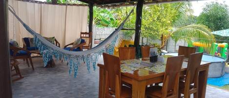 A covered patio with a wooden table and chairs, a hammock, and a pool area.