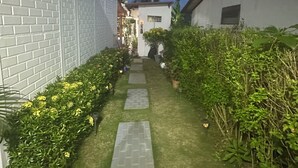Un étroit sentier bordé de murs en briques blanches et de haies verdoyantes, qui mène à un bâtiment surmonté d’une antenne parabolique.