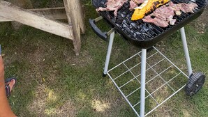 Un barbecue avec de la viande et des légumes, une table en bois avec un saladier et des ustensiles, et un mur de pierre agrémenté de verdure.