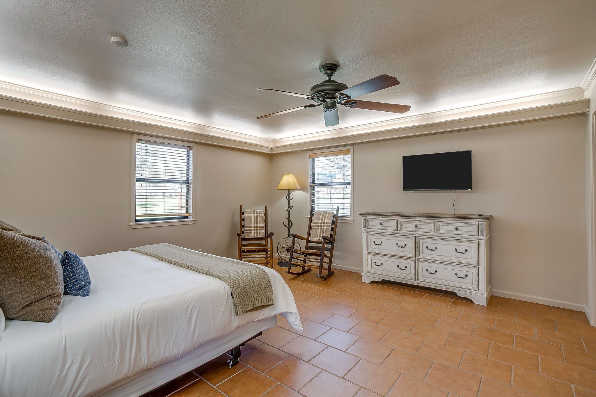 A bedroom with a bed, dresser, two rocking chairs, a ceiling fan, and a TV mounted on the wall.