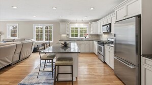 A modern kitchen with a central island, stainless steel appliances, and a dining area with a sofa and chairs. - Hoppy Place: 066-M Expansive Deck area, Hot Tub, Walk to Salt Water (Mashpee)