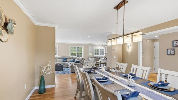 A dining area with a long table set for six, a chandelier, and a view into the living room. - Hoppy Place: 066-M Expansive Deck area, Hot Tub, Walk to Salt Water (Mashpee)