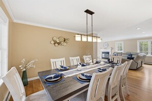 Comedor con una mesa larga dispuesta para seis, sillas blancas, una lámpara moderna y un adorno en la pared.
