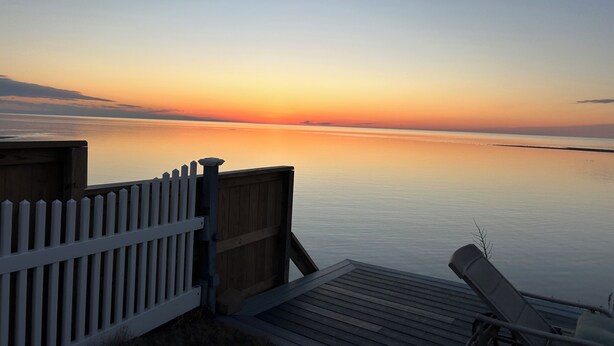 A wooden deck with a white fence and a lounge chair, overlooking a calm body of water during sunset. - Smith Beach cottage w/ private sunset (Cape Charles)