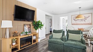 A modern living room with a flat-screen TV, wooden entertainment unit, green sofa, and a dining area with chairs and a table. - Family Arcade Home Pets OK Near Red Rocks (Englewood)
