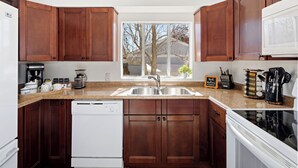 A kitchen with wooden cabinets, a dishwasher, a sink, and a window with a view of trees and a house. - Family Arcade Home Pets OK Near Red Rocks (Englewood)