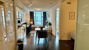 A modern apartment with a kitchen, dining area, and living room. It features dark wooden floors, white cabinetry, and a large window providing natural light.