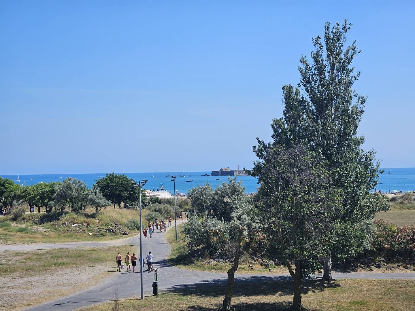 A coastal path with people walking, trees, and a clear view of the sea and boats.